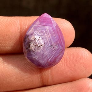 May include: A faceted, teardrop-shaped gemstone with a vibrant pink and purple hue. The gem features a complex internal pattern of lines and a dark, speckled inclusion near the base. The stone is held between fingers, showcasing its polished surface.