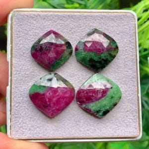 Può includere: Quattro pietre preziose sfaccettate a forma di diamante disposte in formazione quadrata su un cuscinetto espositivo grigio chiaro testurizzato. Ogni pietra presenta una combinazione di colori distinta di magenta intenso e verde bosco.