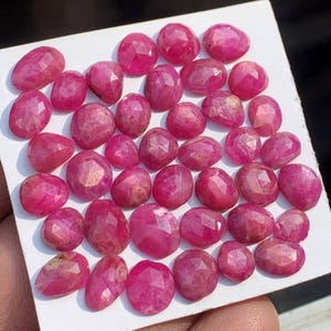 May include: A collection of faceted, oval-shaped ruby gemstones in varying sizes. The rubies display a deep, vibrant pink-red hue and are arranged on a white surface. The gemstones have a polished, reflective surface.