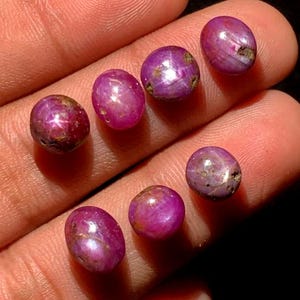 Peut inclure: Collection de sept pierres précieuses rubis ovales polies. Les gemmes sont d'un rouge-rose profond, avec un effet en forme d'étoile visible à la surface. Les pierres précieuses sont tenues dans la main d'une personne.