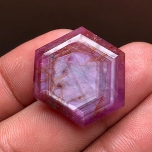 May include: A hexagonal-cut gemstone with a translucent pink and purple body color. The stone features visible internal needle-like inclusions and a distinct geometric pattern with layered bands of color radiating from the center to the edges.