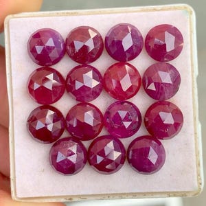 May include: Fourteen round-cut rubies with a faceted surface, displayed in a white tray. The gemstones are a deep red hue, with multiple facets catching the light. The arrangement highlights the rubies' colour and clarity, showcasing their brilliance.