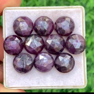 Puede incluir: Nueve piedras preciosas redondas, facetadas, de color púrpura oscuro están dispuestas en una bandeja blanca. Las gemas tienen una superficie pulida y reflectante, y son de un rico color púrpura oscuro. Las gemas son probablemente para la fabricación de joyas.