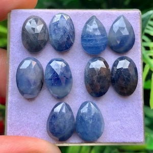 Könnte beinhalten: Eine Sammlung von zehn facettierten blauen Saphiredelsteinen, die auf einem hellvioletten Display-Pad angeordnet sind. Die Steine haben verschiedene Formen, darunter Oval- und Tropfenschliffe, mit tiefblauen Farbtönen und geometrischen Facetten.