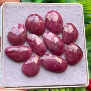 Può includere: Una collezione di dieci pietre preziose sfaccettate di colore rosso rosato intenso disposte su un cuscinetto espositivo grigio chiaro testurizzato. Ogni pietra presenta una superficie lucida a faccette multiple che riflette la luce.
