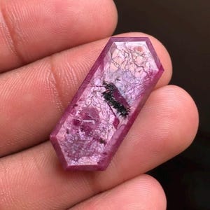 Peut inclure: Une pierre précieuse hexagonale allongée et facettée d'une teinte magenta-rose profonde. La pierre présente une structure cristalline translucide avec des fractures internes visibles et une inclusion sombre en forme d'aiguille au centre.