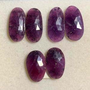 Op de afbeelding: Zeven ovaalvormige, gefacetteerde edelstenen in diepe paarse en magentatinten. De edelstenen hebben een gepolijst, reflecterend oppervlak met zichtbare insluitsels. De opstelling is in twee rijen, met drie edelstenen in de bovenste rij en twee in de onderste rij.