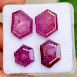 May include: Four faceted, hexagonal, deep red gemstones are displayed in a white plastic case. The gems have a polished appearance with internal patterns. The gems are a rich, saturated colour.