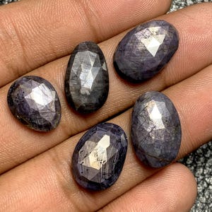 Puede incluir: Cinco piedras preciosas facetadas de forma ovalada en diferentes tonos de azul oscuro y gris. Las piedras preciosas tienen una superficie pulida y reflectante, con estructuras internas visibles. Las piedras se muestran sobre una superficie marrón claro.