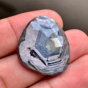 Peut inclure: Une pierre précieuse saphir bleu de forme triangulaire à facettes, présentant des zones internes visibles et des inclusions naturelles. La pierre affiche un mélange de tons bleu profond et gris clair avec une surface polie réfléchissant la lumière.