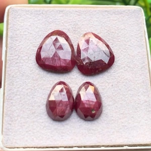 Puede incluir: Cuatro rubíes de color rojo intenso con facetas dispuestos en forma cuadrada. Las dos piedras superiores son más grandes y triangulares, mientras que las dos inferiores son más pequeñas y tienen forma de pera. Cada gema tiene una superficie pulida.