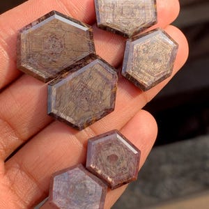 Può includere: Una collezione di sei gemme esagonali con finitura lucida. Le pietre mostrano una miscela di tonalità marroni e viola con motivi lineari interni. Le sfaccettature catturano la luce, creando un effetto scintillante. Le gemme sono tenute in una mano.