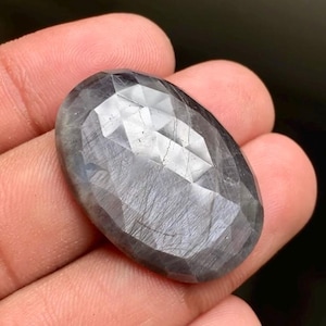 May include: An oval-shaped, faceted gemstone with a dark gray, translucent appearance. The stone features a polished surface with multiple triangular facets that reflect light, showcasing internal linear inclusions and a smooth, rounded edge.