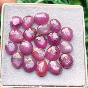 Pode incluir: Uma coleção de dezassete rubis cor-de-rosa facetados de forma oval dispostos numa almofada de exposição cinzenta clara texturada. Cada pedra preciosa apresenta uma superfície polida com múltiplas facetas triangulares.