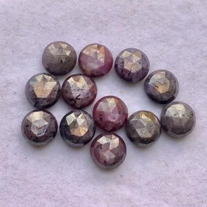 May include: A selection of faceted, round gemstones in shades of purple, pink, and grey. The gemstones have a geometric cut, reflecting light. The arrangement is scattered on a light-coloured surface, showcasing the gems' varied hues and textures.