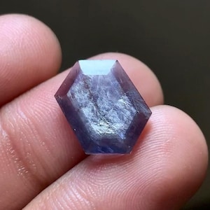 Puede incluir: Una gema hexagonal facetada, de color púrpura intenso. La gema tiene una superficie pulida con inclusiones internas en forma de nube y se sostiene entre los dedos. La forma de la gema es simétrica, con bordes definidos.