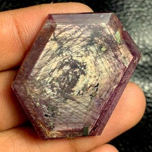 May include: A faceted, hexagonal gemstone with a deep purple hue. The stone features internal patterns of black and white, resembling a network of fine lines. Small flecks of green and gold are visible within the gem, adding to its unique appearance.