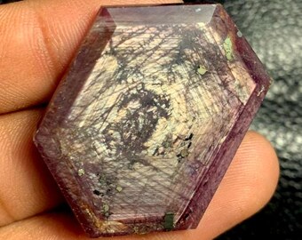 Puede incluir: Una gema facetada, hexagonal, de color púrpura intenso. La piedra presenta patrones internos en blanco y negro, que se asemejan a una red de finas líneas. Se ven pequeñas motas verdes y doradas dentro de la gema.