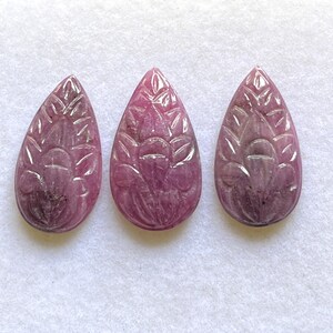 Peut inclure: Trois pierres précieuses taillées en forme de goutte d'eau, de différentes nuances de rose et de violet. Chaque pierre présente un motif floral détaillé. Les pierres sont exposées sur une surface blanche, mettant en valeur leurs gravures et variations de couleurs.