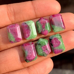 Può includere: Sette gemme sfaccettate con sfumature rosa e verdi. Le gemme hanno forme rettangolari ed esagonali, con un aspetto lucido e traslucido. Le gemme sono tenute in una mano.