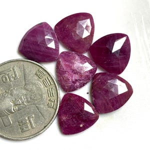 May include: Six faceted ruby gemstones in a triangular shape. The gemstones are a deep pink colour and have a slight sparkle. The gemstones are arranged in a circle around a Korean coin.