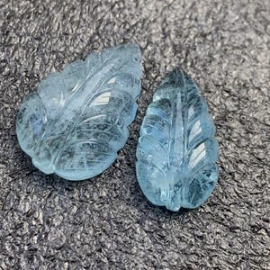 May include: Two aquamarine gemstone leaf carvings. The leaves are intricately carved with detailed veining, showcasing a light blue color. The gemstones are displayed on a textured, dark gray surface.