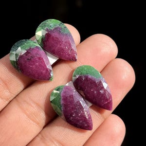May include: Four faceted, teardrop-shaped gemstones. The gems have a deep magenta hue at the base, transitioning to a vibrant green at the top. The gems are displayed on a light surface, with a dark background.