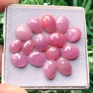 Pode incluir: Uma coleção de catorze pedras preciosas cor-de-rosa facetadas dispostas num expositor quadrado de feltro cinzento claro. Cada pedra apresenta uma superfície polida com múltiplas facetas que reflete a luz, exibindo um tom rosa vibrante.