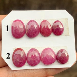 May include: Six oval-shaped, faceted, deep red gemstones are arranged in two rows of three on a white card. The gemstones have a polished, reflective surface, and are numbered 1 and 2.
