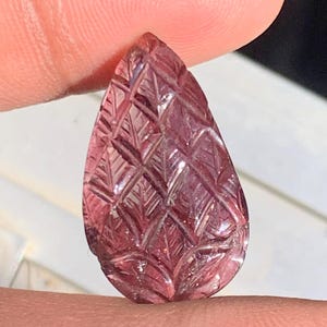 May include: A teardrop-shaped, carved gemstone in shades of pink and purple. The gemstone features a detailed pattern of intersecting lines and leaf-like shapes, creating a textured surface. The gem is held between two fingers.