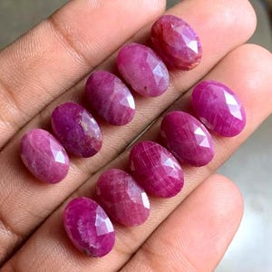 Puede incluir: Una colección de piedras preciosas de rubí ovaladas y facetadas en varios tonos de rojo y rosa. Las piedras se muestran sobre una superficie clara, mostrando sus facetas pulidas y patrones internos. Ideales para la creación de joyas.