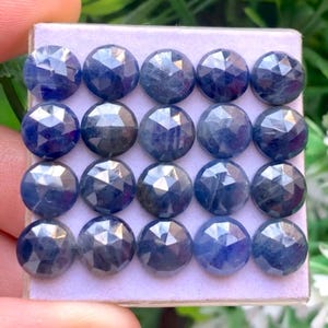 Può includere: Una collezione di venti gemme di zaffiro blu, rotonde e sfaccettate, disposte in una griglia quattro per cinque su un cartoncino espositivo color viola chiaro. Ogni gemma presenta un taglio a rosa simmetrico con molte sfaccettature.