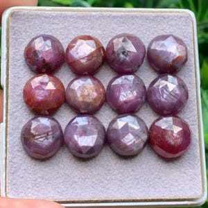 Puede incluir: Una colección de doce piedras preciosas redondas y facetadas de color rosa intenso y rojo, dispuestas en una cuadrícula de tres por cuatro. Cada piedra presenta una superficie pulida de múltiples facetas que refleja la luz.