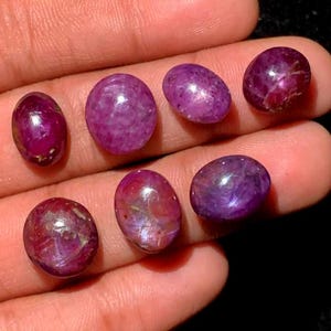 May include: A collection of seven oval-shaped, polished gemstones in varying shades of deep red and purple. The gems are held in a hand, showcasing their smooth surfaces and internal patterns. The stones have a natural, organic appearance.