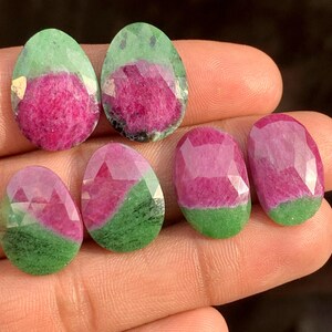 Pode incluir: Uma coleção de pedras preciosas facetadas, mostrando uma vibrante mistura de cores. As pedras apresentam uma combinação marcante de magenta profundo, verde e facetas claras. As pedras preciosas são ovais e em forma de gota, perfeitas para fazer joias.