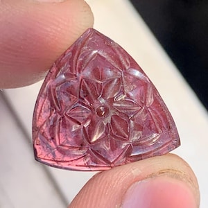 May include: A triangular, faceted gemstone with a floral design carved into its surface. The gem is a deep pink color, and the carving creates a play of light and shadow. The gem is held between two fingers.