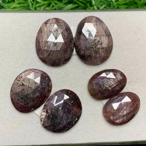 May include: Five oval-shaped, faceted gemstones with a dark purple hue and a shimmery, iridescent finish. The gemstones are arranged on a white surface.