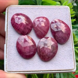 Pode incluir: Cinco pedras preciosas facetadas de um vermelho rosado profundo dispostas numa almofada de exibição cinzenta clara. Cada pedra apresenta uma superfície lapidada em rosa com um padrão geométrico de facetas triangulares que refletem a luz.