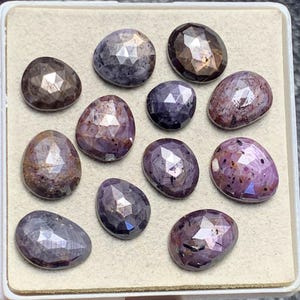 Puede incluir: Una colección de gemas facetadas de forma ovalada en tonos púrpura, marrón y gris. Las gemas están dispuestas en una bandeja rectangular, mostrando sus superficies pulidas y patrones internos. Las piedras varían en color y claridad.