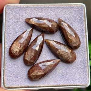 May include: A collection of six faceted, pear-shaped gemstones with a deep reddish-brown hue. Each stone features a polished surface with multiple geometric facets that reflect light, arranged in a cluster on a light purple textured display pad.