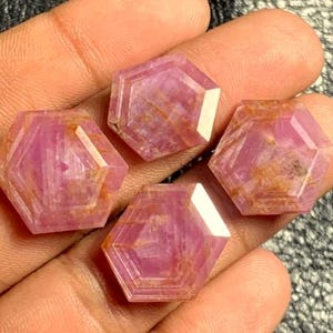 Puede incluir: Cuatro gemas hexagonales facetadas, en tonos rosa y rojo. Las gemas tienen una superficie pulida con patrones internos visibles. Las gemas se sostienen en una mano, mostrando su tamaño y color.