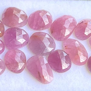 Puede incluir: Un conjunto de gemas rosas facetadas, de forma ovalada. Las piedras, de tamaños variados, presentan una calidad translúcida y patrones internos visibles. Se exhiben sobre una superficie blanca, resaltando su color y talla.