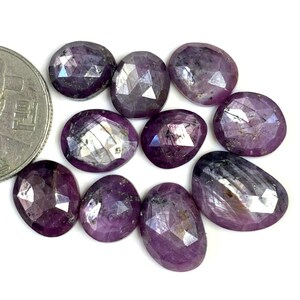 Peut inclure: Dix pierres précieuses de saphir violet facettées, chacune avec une forme et une taille uniques, sont disposées sur une surface blanche. Les pierres précieuses ont un éclat argenté et mesurent environ la taille d'une pièce de dix centimes d'euro.