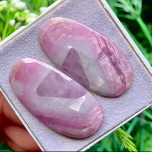 May include: Two oval-shaped, faceted gemstones featuring a smooth, polished surface with a distinct colour gradient transitioning from deep magenta and lavender to translucent white. The stones are set against a light purple, textured fabric lining.