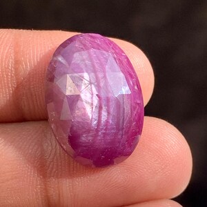 Può includere: Una gemma di rubino ovale e sfaccettata. La gemma è di un colore magenta intenso con riflessi interni. La superficie è tagliata con molteplici sfaccettature, creando un effetto scintillante. La gemma è tenuta in una mano.