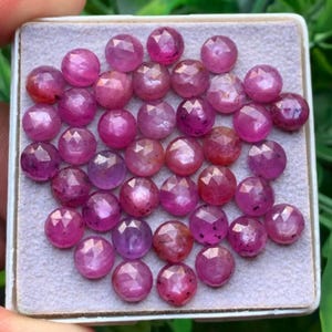 May include: A collection of small, round, faceted pink rubies arranged in a square white display box. Each gemstone features a multi-faceted surface that reflects light, showcasing various shades of deep pink and magenta across the group.