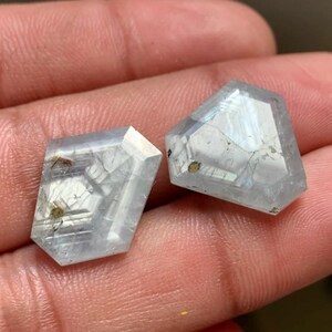 Peut inclure: Deux pierres précieuses taillées en pentagone, d'une teinte bleu pâle translucide. Les pierres présentent des surfaces à facettes, des inclusions internes et des motifs géométriques visibles dans leur structure, avec une finition polie.