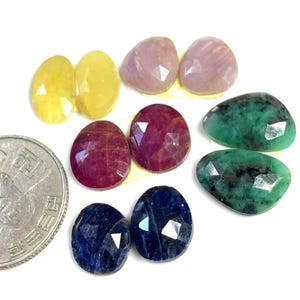 Puede incluir: Surtido de cabujones de piedras preciosas facetadas en varios colores, incluyendo rosa, amarillo, verde, azul y rojo. Las piedras preciosas se muestran sobre una superficie blanca.
