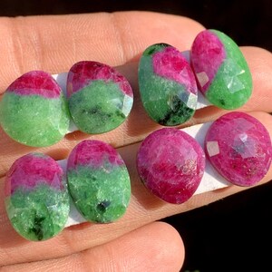 May include: A collection of faceted gemstones, showcasing a vibrant mix of colors. The stones feature a striking combination of deep red and verdant green hues, with some displaying a gradient effect. The gems are oval-shaped and appear to be held on a white surface.