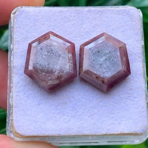 May include: Two loose, hexagonal-cut gemstones with a translucent, pale pinkish-purple hue and visible internal inclusions. The stones are displayed side-by-side on a square, light lavender-coloured foam insert within a clear plastic storage case.
